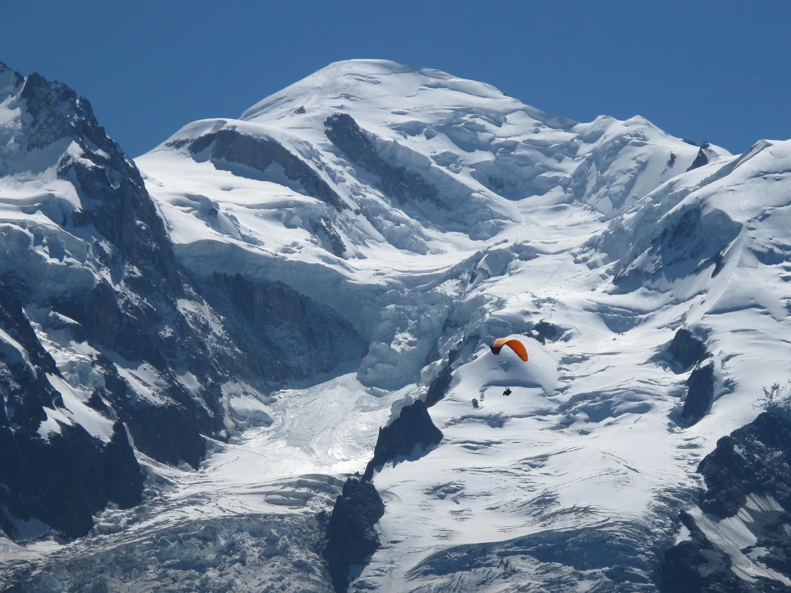 Snow-covered summit of Mont Blanc against a blue sky, seen from Planpraz above Chamonix, with a paraglider in front of the glaciers