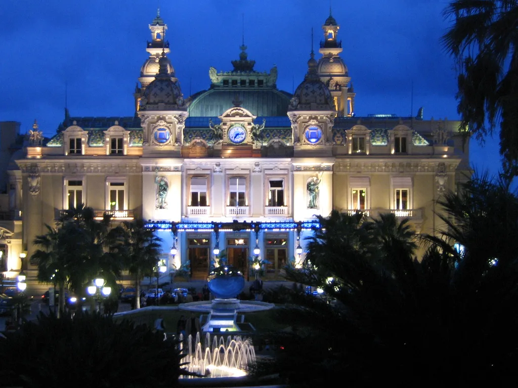 The Casino de Monte-Carlo lit up at dusk, its domed towers and clock against a deep blue sky, with palms and a fountain in front